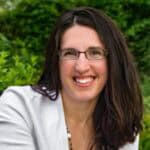 Cropped headshot of Danielle Coleman, owner of Deeper Living Adventures. She has dark hair that drops several inches below her left shoulder, with it flowing back over her right shoulder. She wears black thinly rimmed glasses, a white jacket, and is smiling. Green trees are in the background.