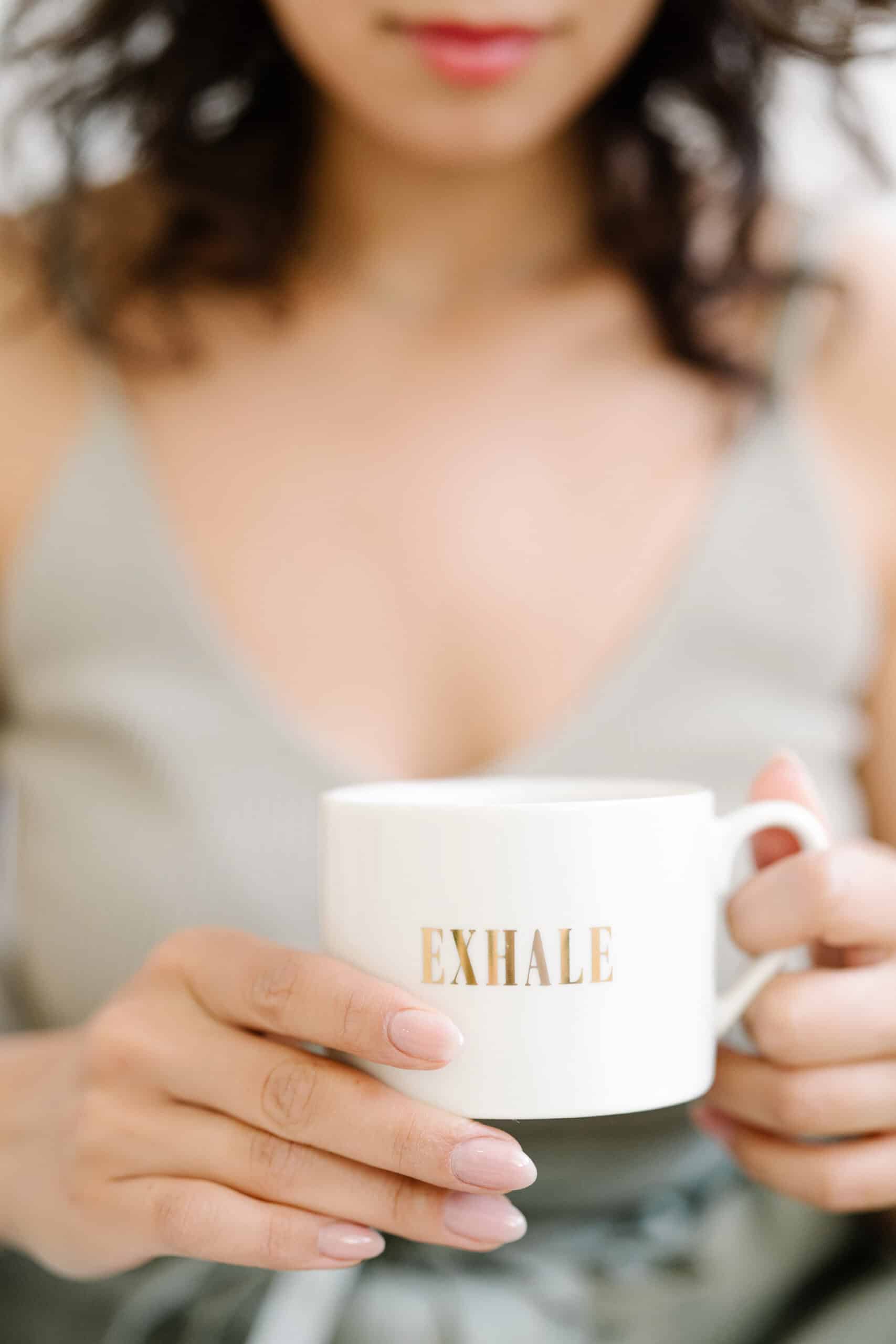 Relaxed woman holding a white mug with "EXHALE" written in gold.